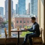 A man sits at a cafe table, working on a laptop and enjoying a city view through large windows