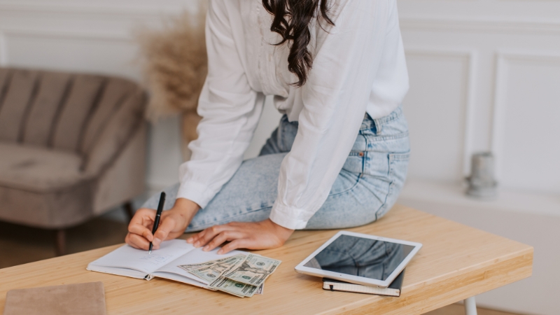 A student in STEM writes budget notes on paper beside cash and a tablet on a desk