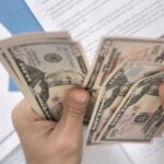 A person counting dollar bills over financial charts, representing small business cash flow management