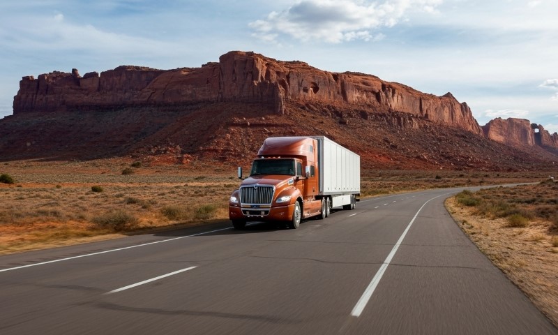 A semi truck drives along a road with a mountain backdrop, showcasing a scenic transportation route