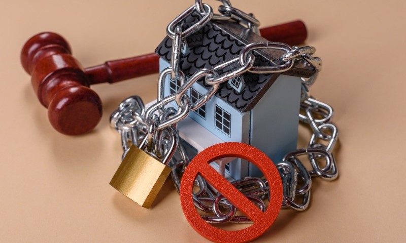 A gavel, a small house model wrapped in chains with a padlock, and a red prohibition sign