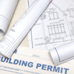 Building permit document surrounded by architectural plans and residential construction drawings.