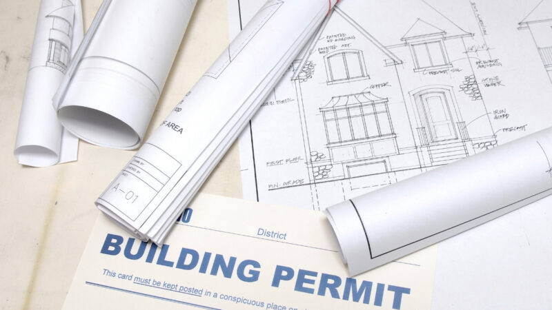 Building permit document surrounded by architectural plans and residential construction drawings.