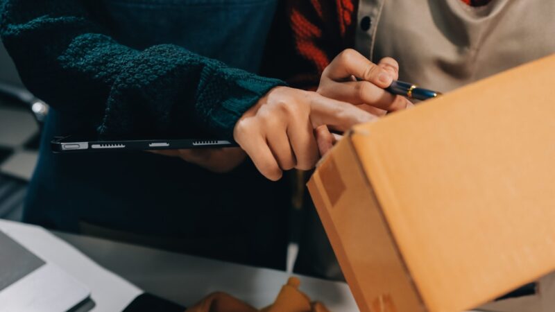 A worker checks a cardboard package beside a tablet.