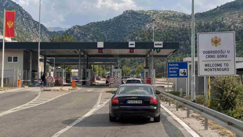 Cars at a Montenegro border checkpoint with a welcome sign and mountains in the background.